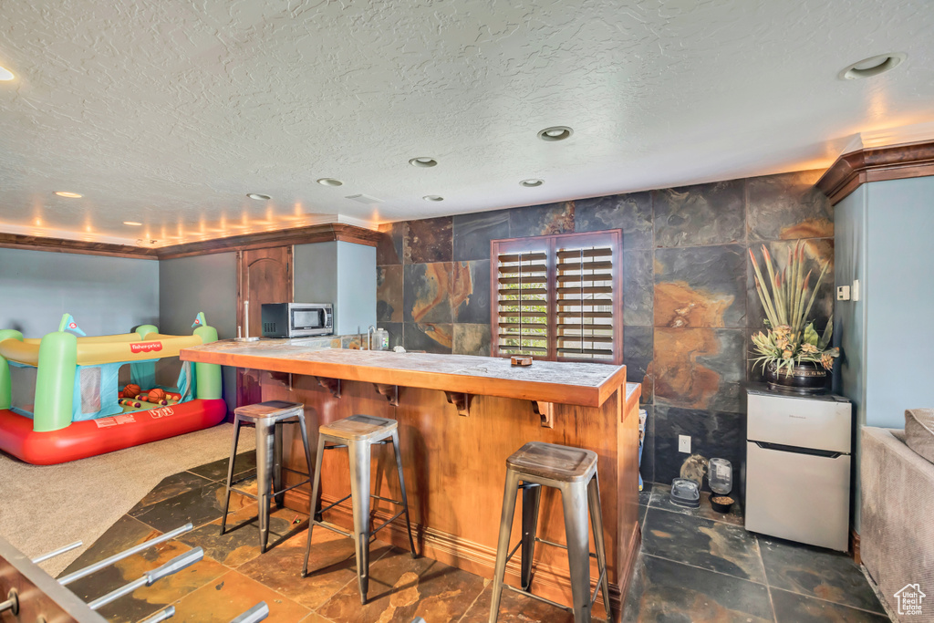 Bar area featuring stainless steel microwave, freestanding refrigerator, a textured ceiling, recessed lighting, and stone finish flooring