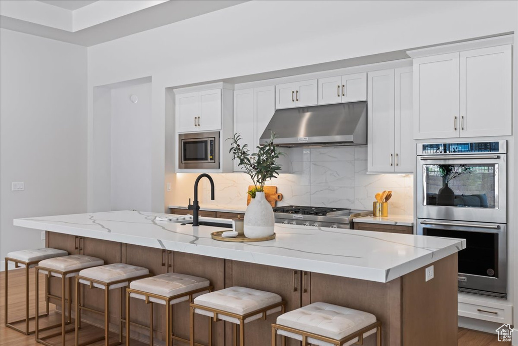 Kitchen featuring under cabinet range hood, appliances with stainless steel finishes, decorative backsplash, white cabinetry, and wood finished floors