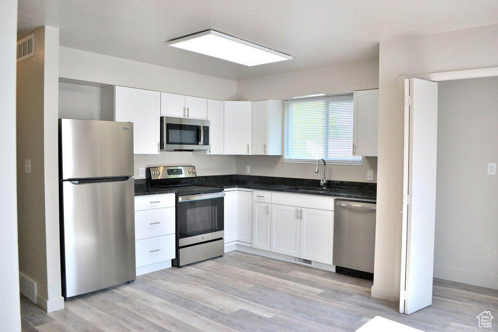 Kitchen with appliances with stainless steel finishes, white cabinets, light wood-style flooring, a sink, and dark stone counters