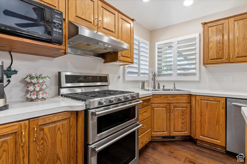 Kitchen with appliances with stainless steel finishes, under cabinet range hood, a sink, dark wood-style floors, and brown cabinetry