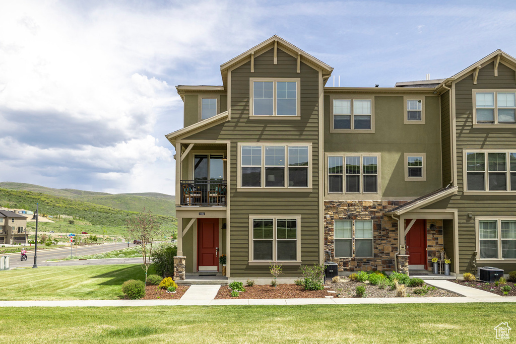 View of front of property featuring a mountain view, stone siding, a balcony, a front lawn, and central air condition unit