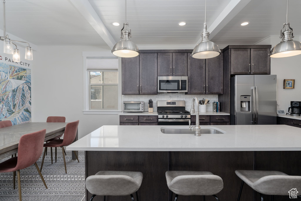 Kitchen featuring stainless steel appliances, a sink, beamed ceiling, a kitchen bar, and light countertops