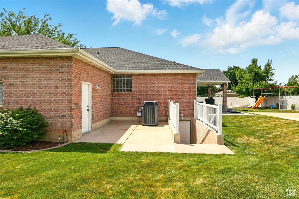 Rear view of house with central AC unit, roof with shingles, a playground, a yard, and a patio area