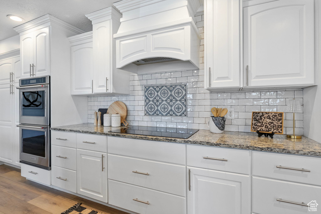 Kitchen with custom exhaust hood, stainless steel double oven, black electric stovetop, wood finished floors, and stone counters