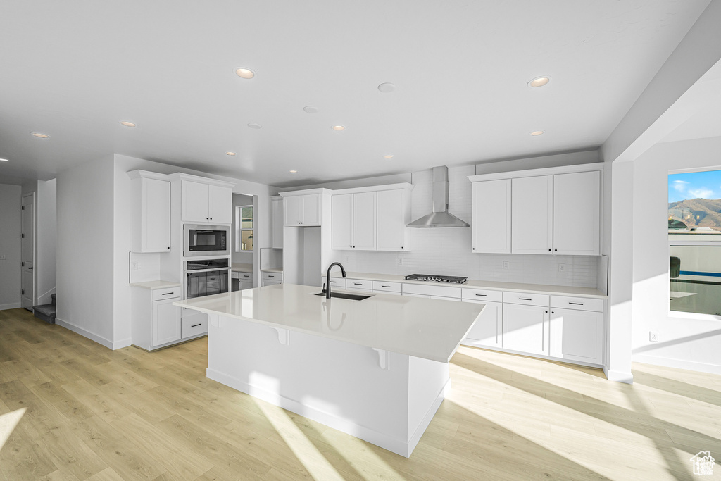 Kitchen with wall chimney range hood, a sink, built in microwave, backsplash, and light wood-style floors