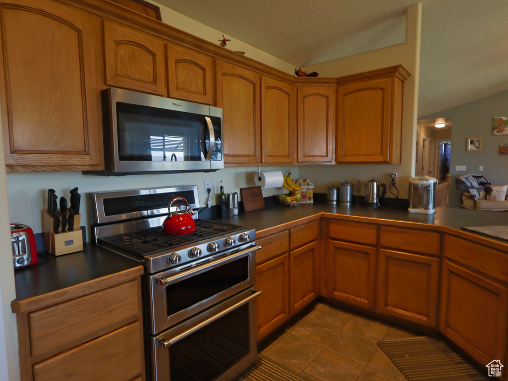Kitchen featuring stainless steel appliances, vaulted ceiling, dark countertops, brown cabinetry, and a textured ceiling