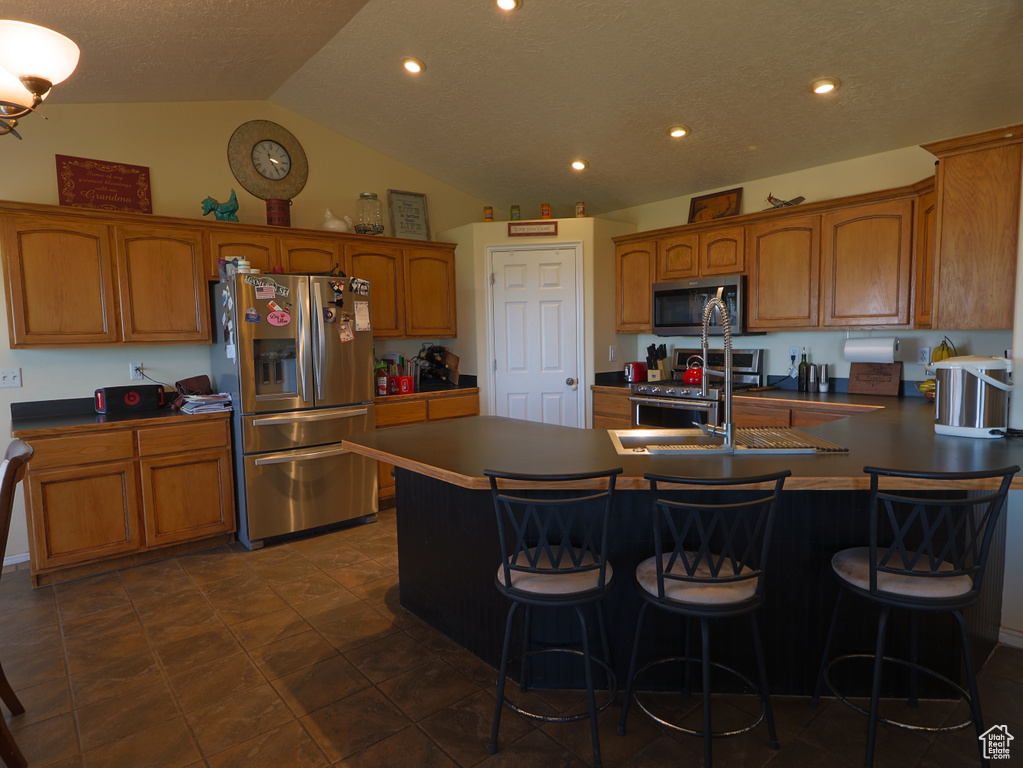 Kitchen featuring appliances with stainless steel finishes, brown cabinetry, a sink, vaulted ceiling, and a kitchen breakfast bar
