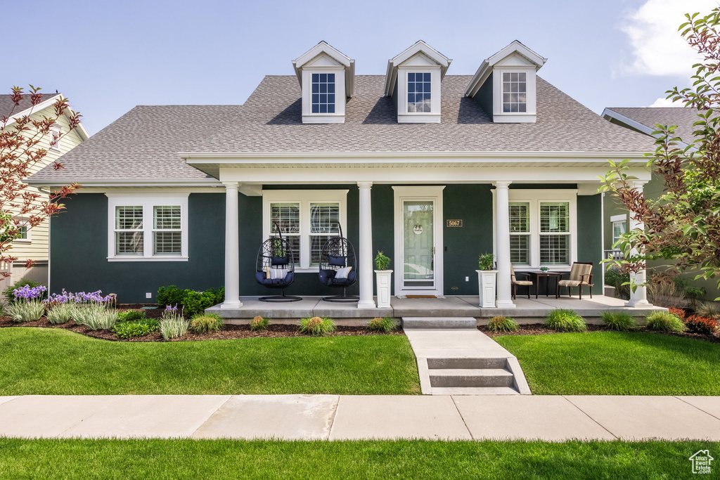 New england style home with a porch, a shingled roof, a front yard, and stucco siding