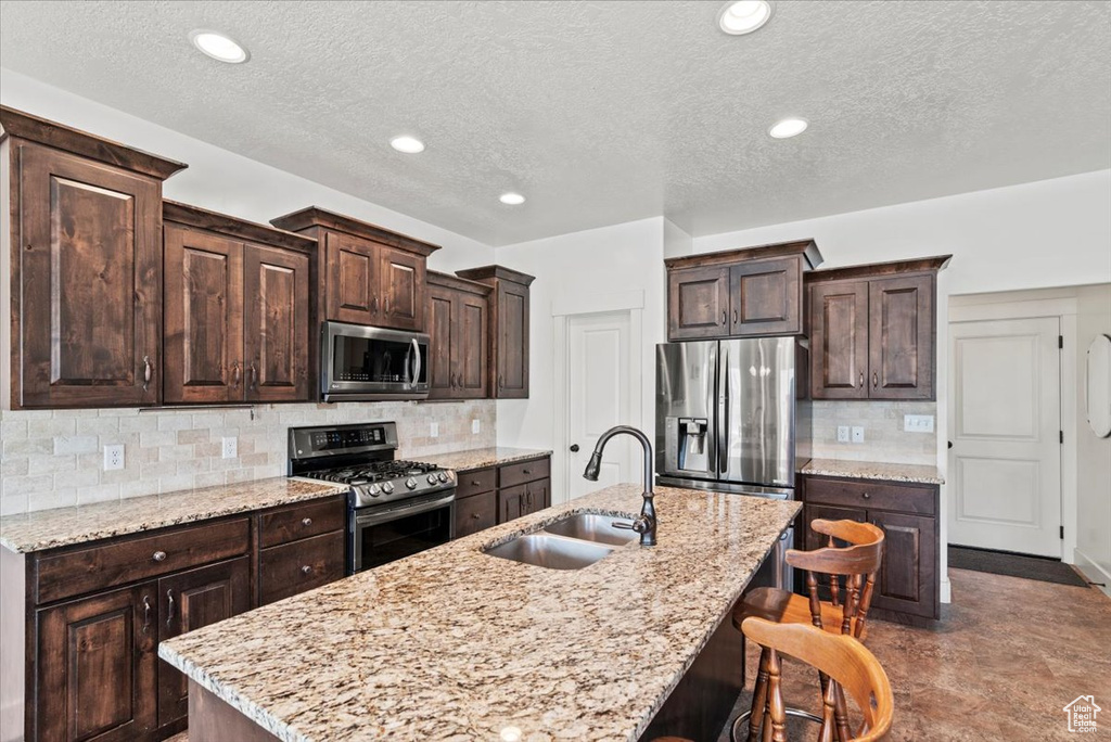 Kitchen featuring appliances with stainless steel finishes, a sink, dark brown cabinetry, tasteful backsplash, and an island with sink