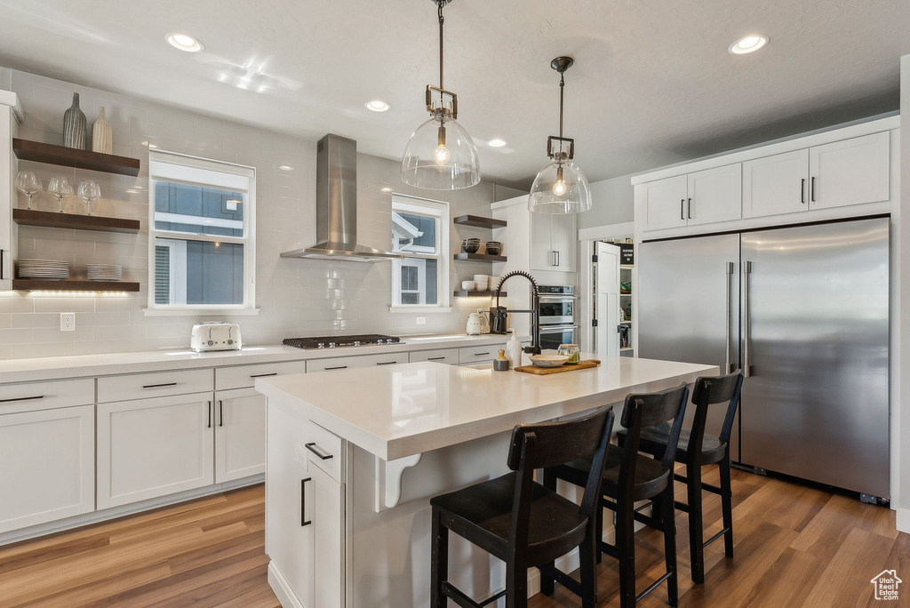 Kitchen featuring appliances with stainless steel finishes, wall chimney range hood, open shelves, tasteful backsplash, and light wood-type flooring