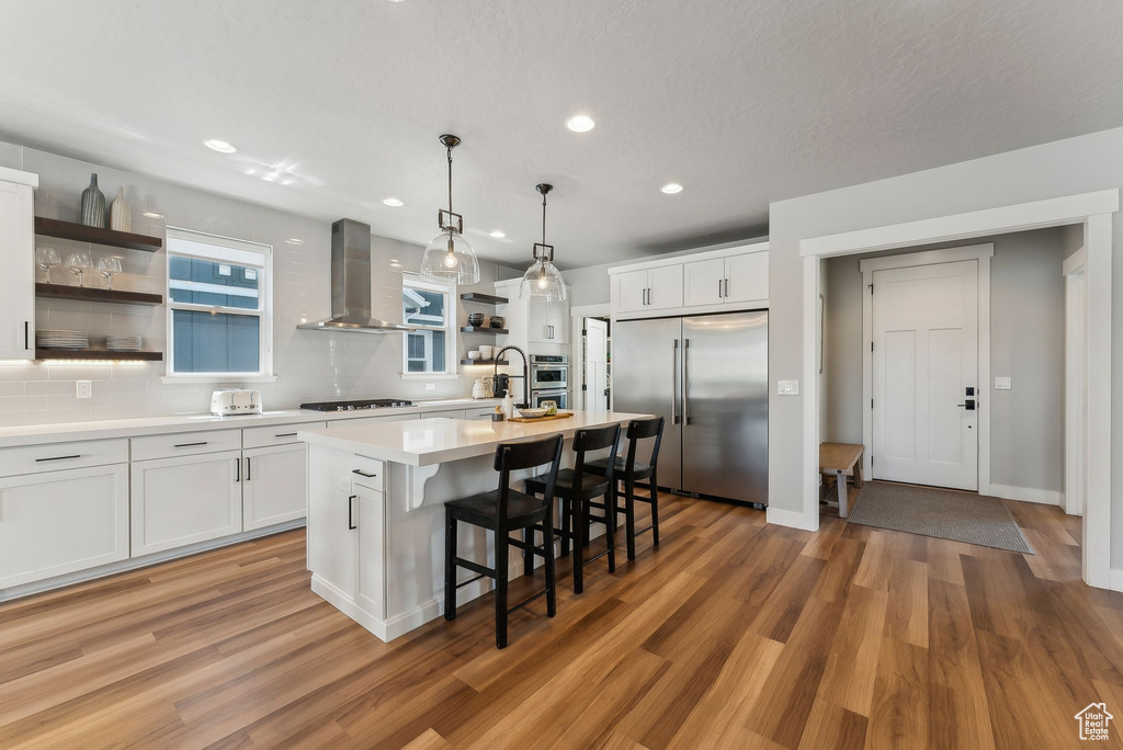 Kitchen featuring appliances with stainless steel finishes, open shelves, light countertops, wall chimney range hood, and a kitchen bar