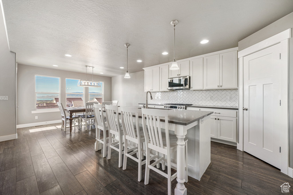 Kitchen featuring stainless steel microwave, backsplash, baseboards, an island with sink, and dark wood-style floors