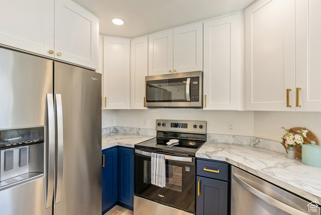 Kitchen featuring appliances with stainless steel finishes, white cabinetry, blue cabinetry, light stone counters, and recessed lighting