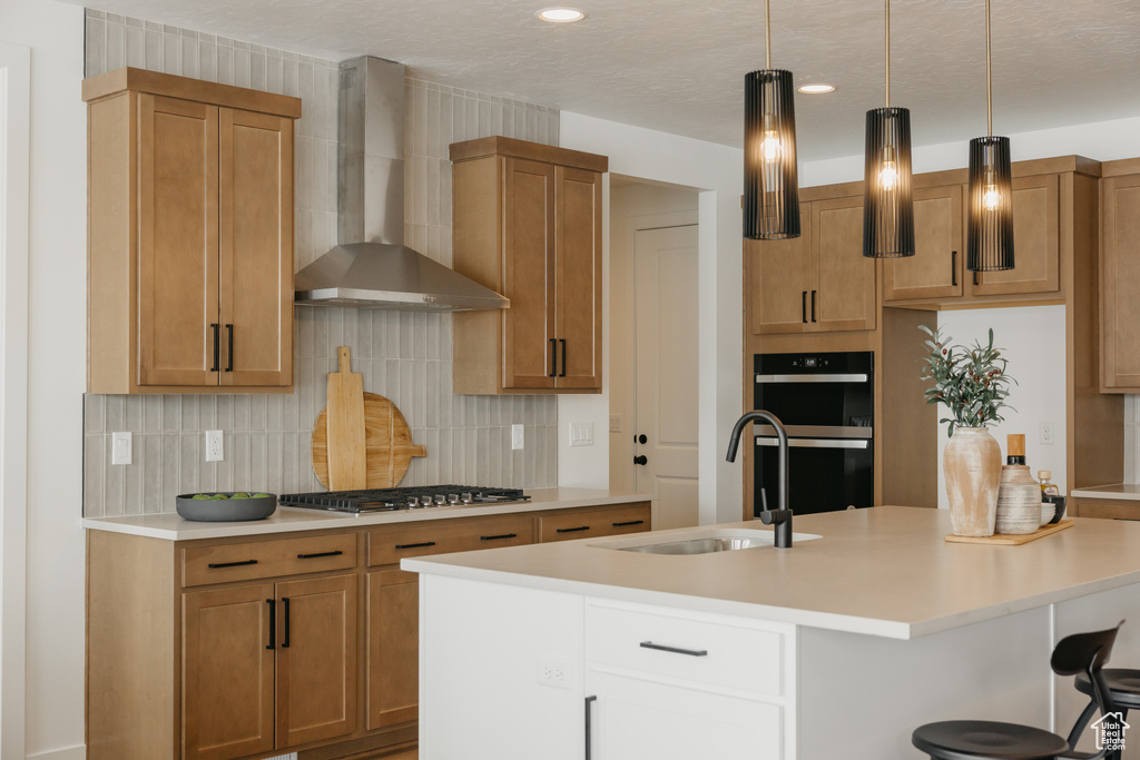 Kitchen with wall chimney exhaust hood, stainless steel gas cooktop, a sink, tasteful backsplash, and a center island with sink