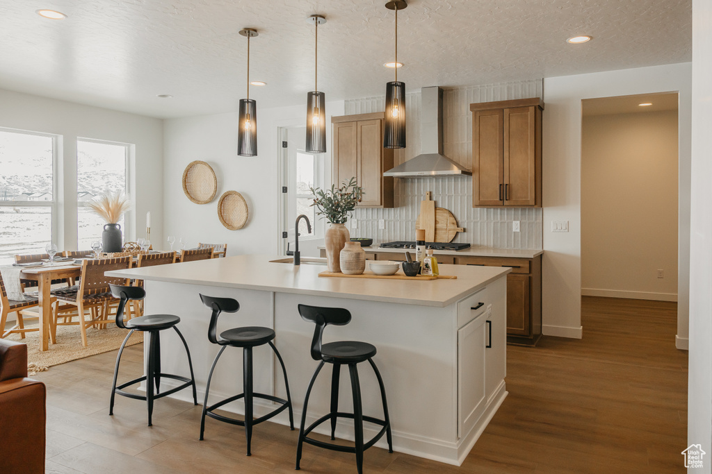 Kitchen with wall chimney exhaust hood, a breakfast bar area, tasteful backsplash, light countertops, and an island with sink
