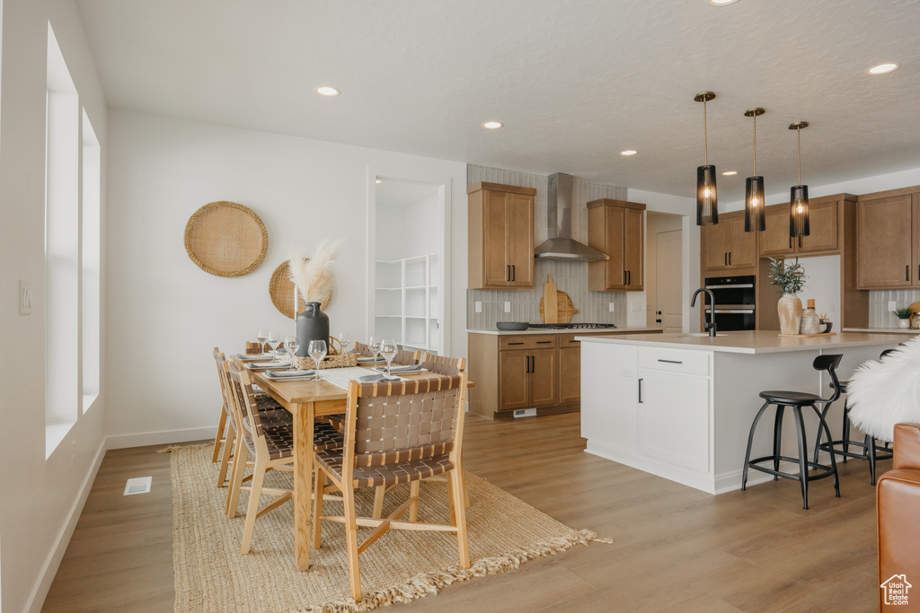 Kitchen with wall chimney range hood, decorative backsplash, light wood-style flooring, double wall oven, and a kitchen bar