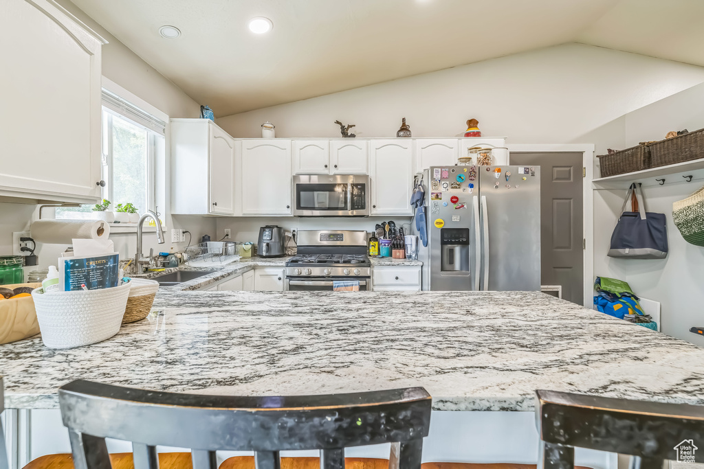 Kitchen featuring stainless steel appliances, a sink, light stone counters, vaulted ceiling, and white cabinetry