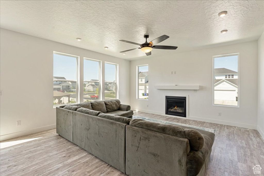 Living room featuring light wood finished floors, a glass covered fireplace, ceiling fan, baseboards, and a textured ceiling
