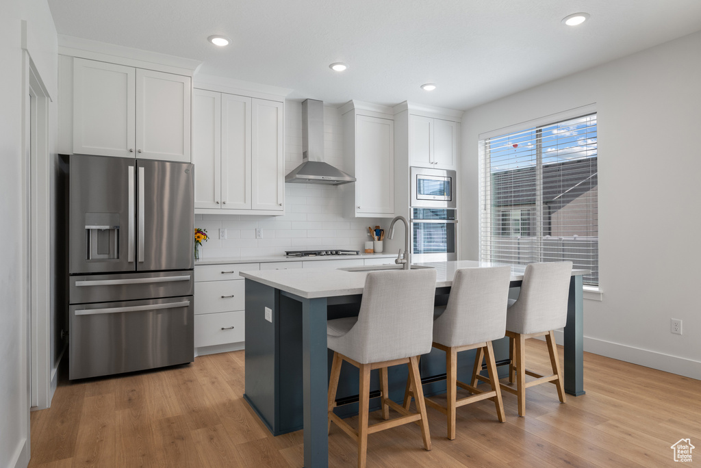 Kitchen with stainless steel appliances, wall chimney range hood, a sink, a kitchen island with sink, and light countertops