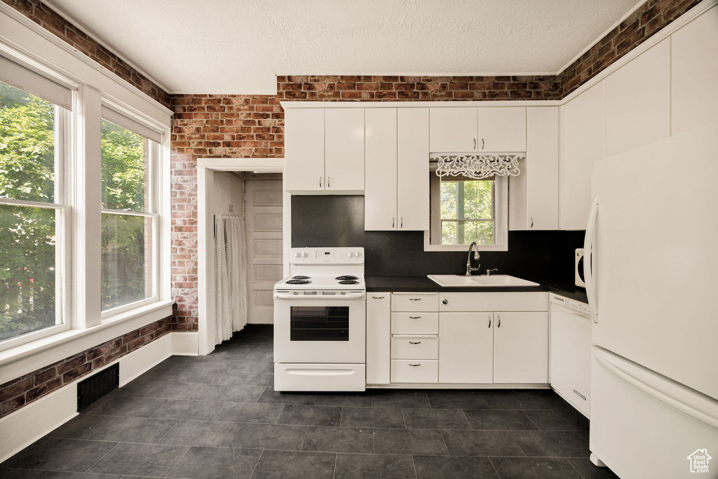 Kitchen with white appliances, a sink, dark countertops, white cabinetry, and brick wall