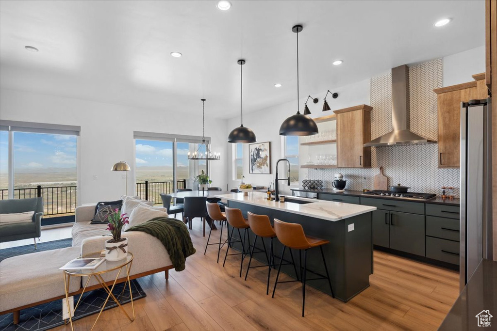 Kitchen with wall chimney exhaust hood, a sink, a kitchen breakfast bar, light wood-style floors, and an island with sink