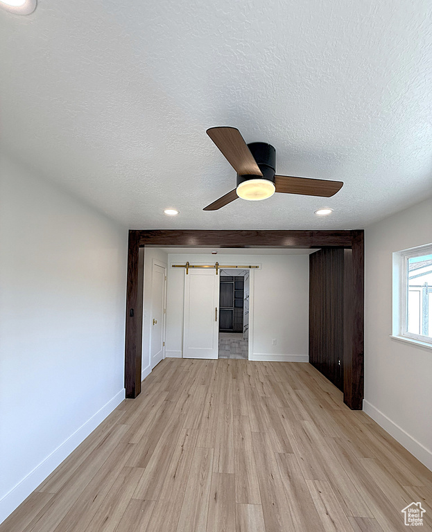 Unfurnished bedroom featuring a barn door, light wood-style flooring, a textured ceiling, baseboards, and ceiling fan
