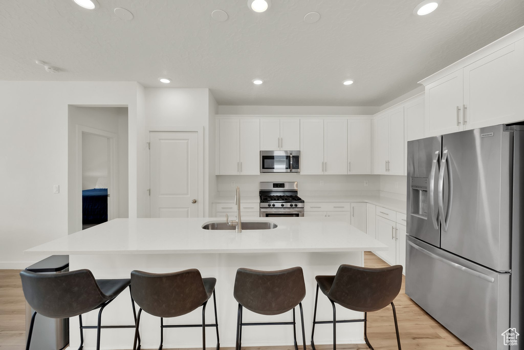 Kitchen with stainless steel appliances, a sink, a kitchen island with sink, light wood-style flooring, and white cabinets