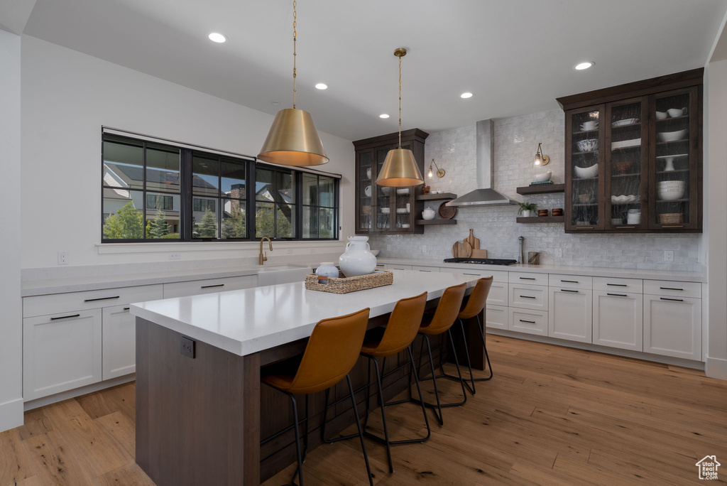 Kitchen featuring wall chimney range hood, light countertops, backsplash, a kitchen island, and glass insert cabinets