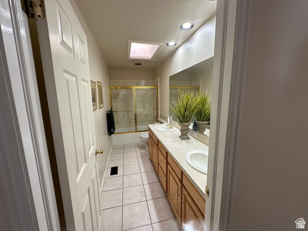 Bathroom with a skylight, double vanity, bath / shower combo with glass door, toilet, and tile patterned flooring