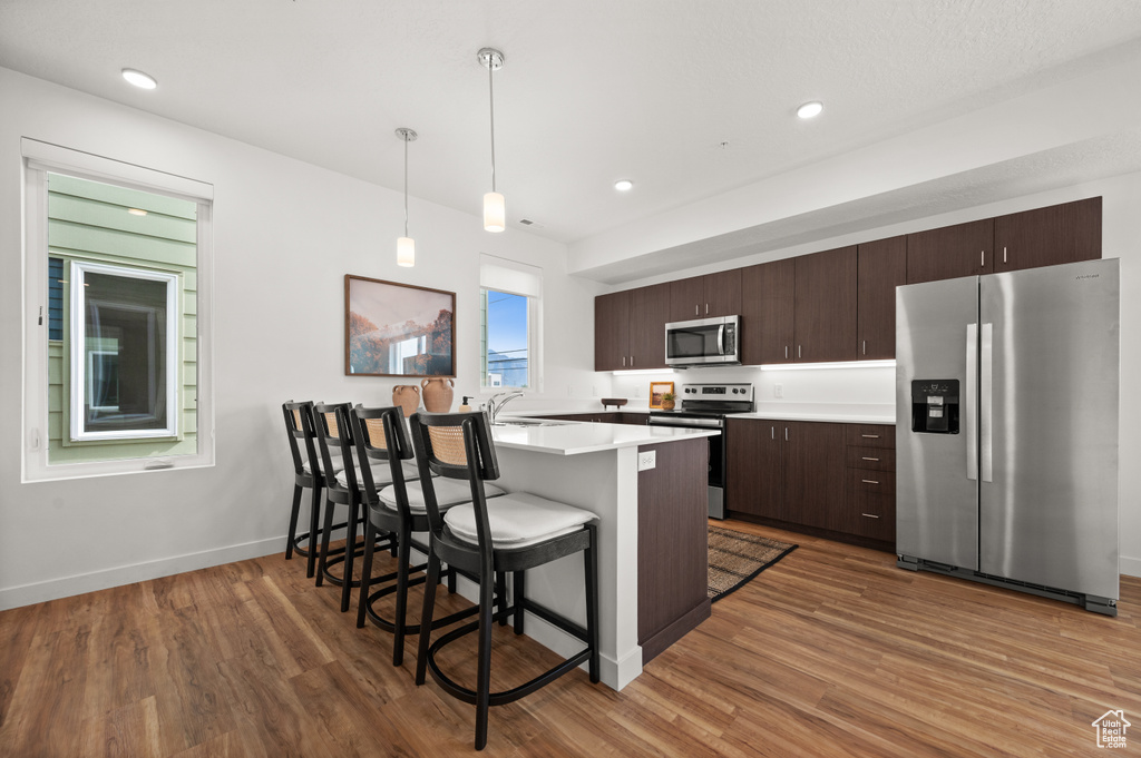 Kitchen featuring appliances with stainless steel finishes, a kitchen breakfast bar, dark brown cabinetry, wood finished floors, and recessed lighting