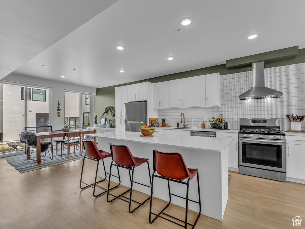 Kitchen featuring appliances with stainless steel finishes, wall chimney range hood, light countertops, a breakfast bar, and white cabinetry