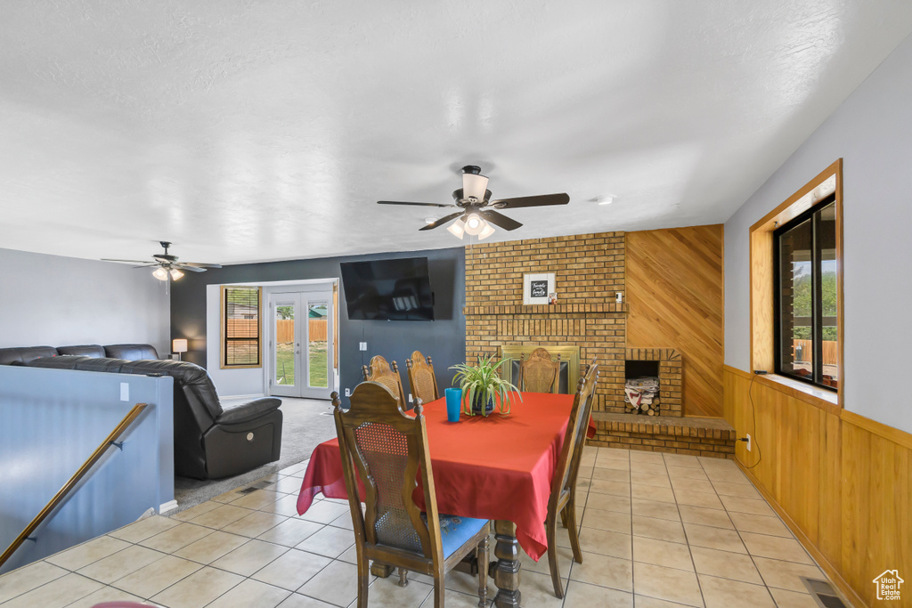 Dining room featuring ceiling fan, wooden walls, light tile patterned floors, french doors, and a brick fireplace