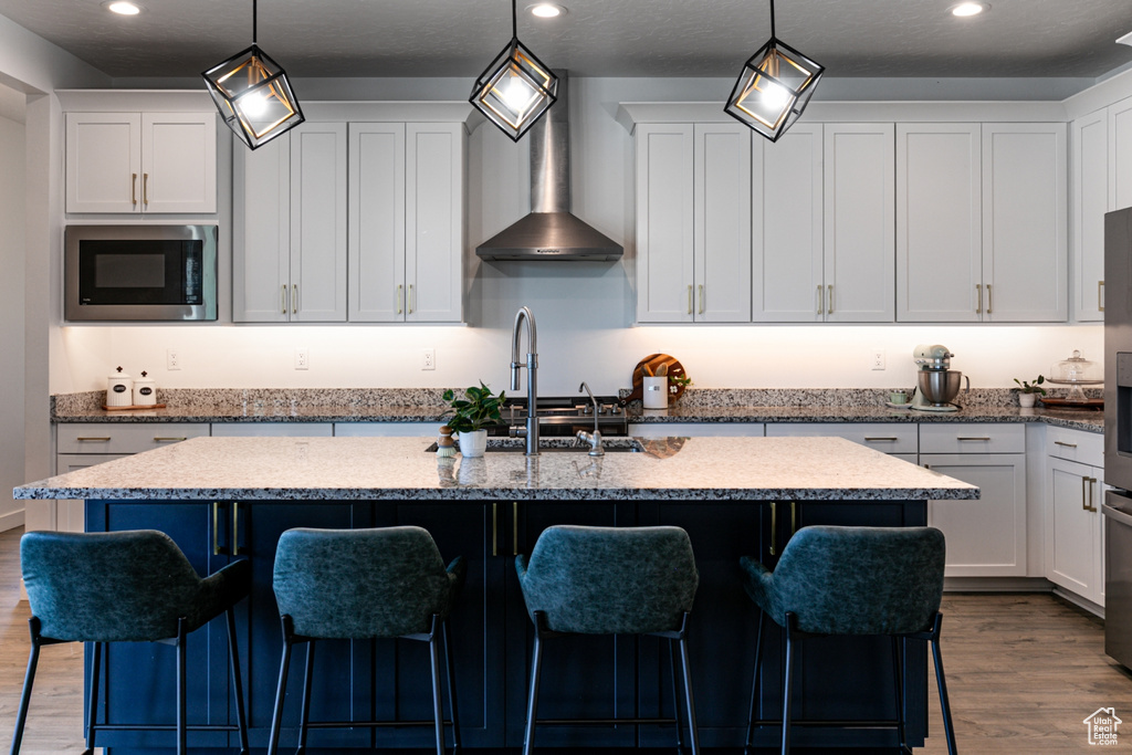 Kitchen featuring stainless steel microwave, wall chimney range hood, wood finished floors, an island with sink, and white cabinets