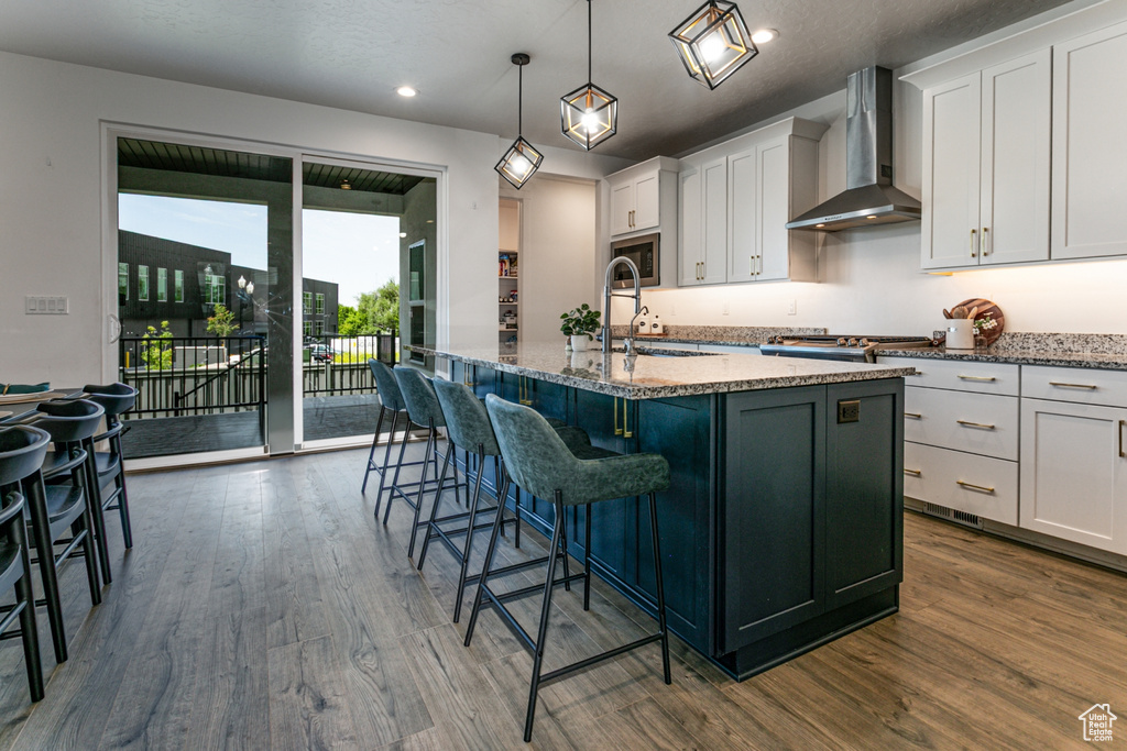 Kitchen featuring wall chimney exhaust hood, an island with sink, dark wood-style flooring, white cabinets, and stone countertops