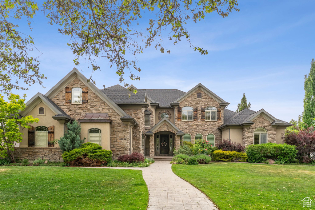 View of front facade with stone siding, a front yard, stucco siding, and roof with shingles