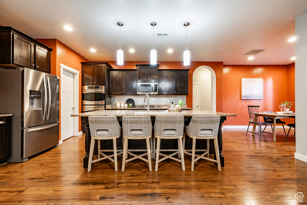 Kitchen with appliances with stainless steel finishes, arched walkways, a kitchen island with sink, tasteful backsplash, and dark wood-style flooring