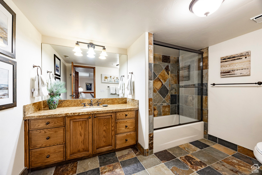 Full bathroom with stone tile floors, baseboards, vanity, toilet, and combined bath / shower with glass door