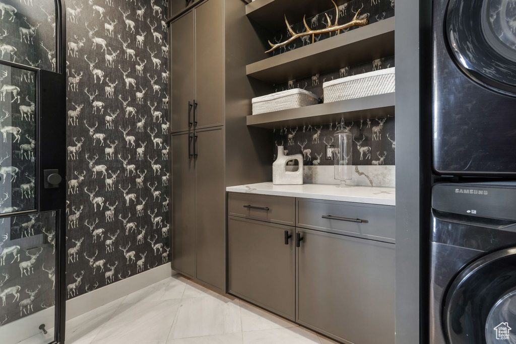 Laundry room with stacked washing machine and dryer, wallpapered walls, and light marble finish flooring