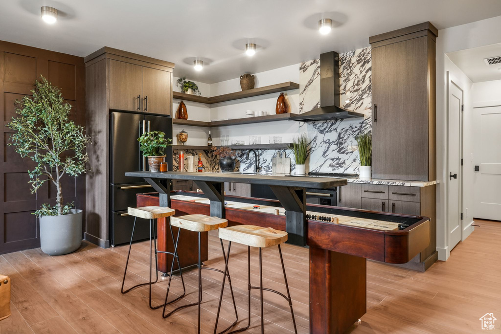 Kitchen featuring wall chimney exhaust hood, black fridge, open shelves, light wood-type flooring, and light countertops