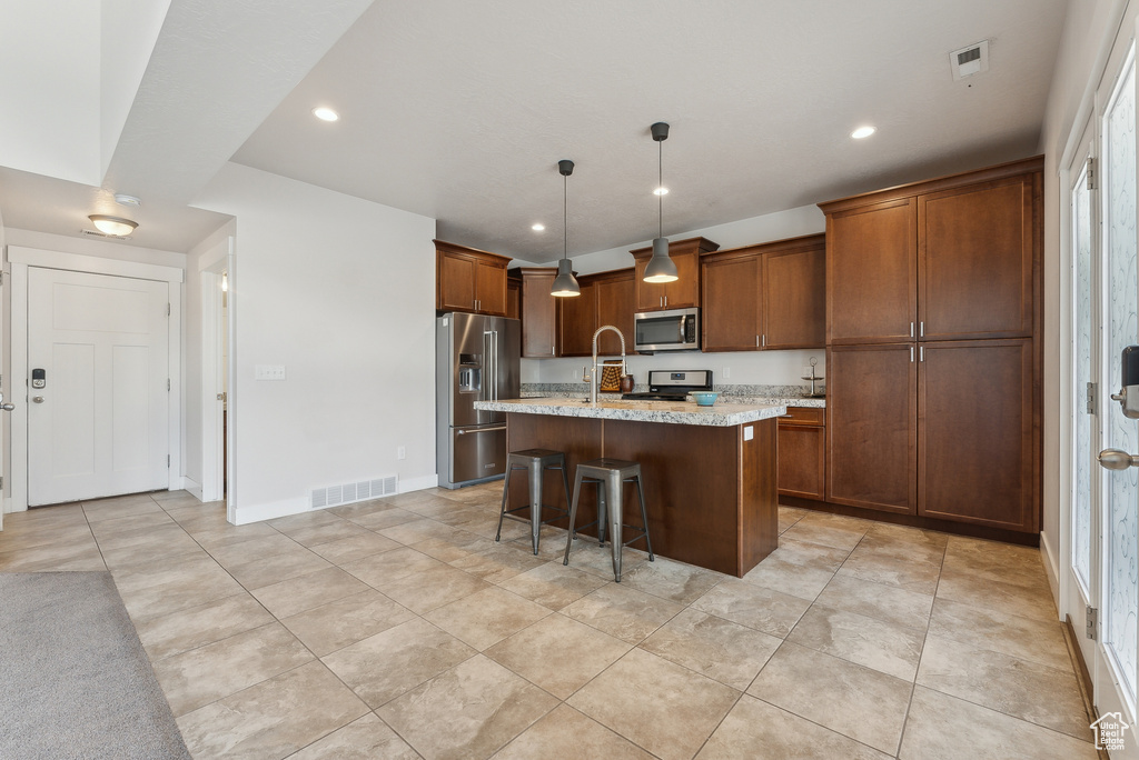 Kitchen with stainless steel appliances, a kitchen island with sink, light tile patterned floors, brown cabinets, and a breakfast bar