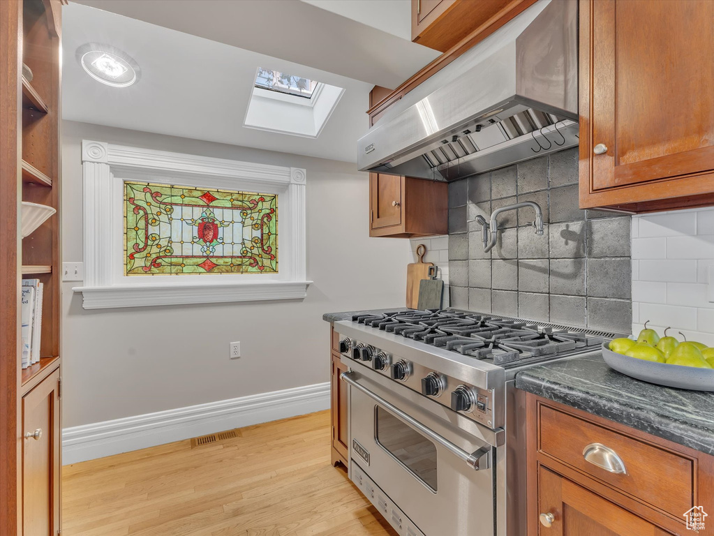 Kitchen featuring luxury stove, wall chimney exhaust hood, a skylight, brown cabinets, and backsplash