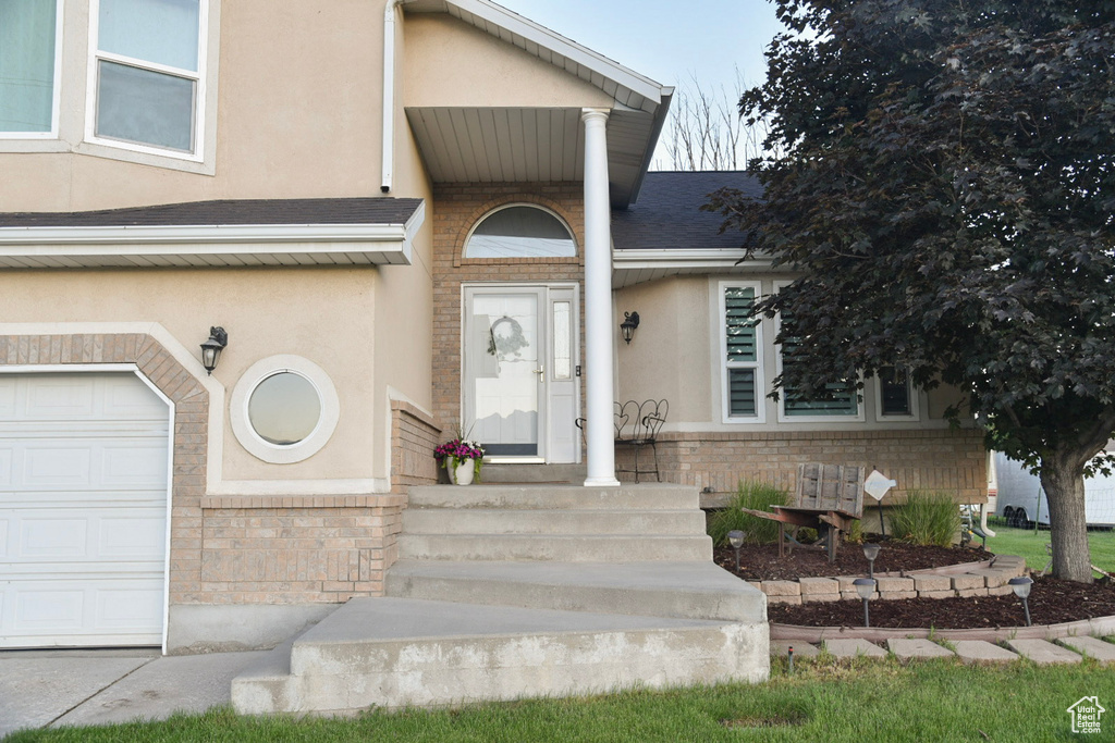 View of exterior entry featuring stucco siding, a garage, brick siding, and a shingled roof