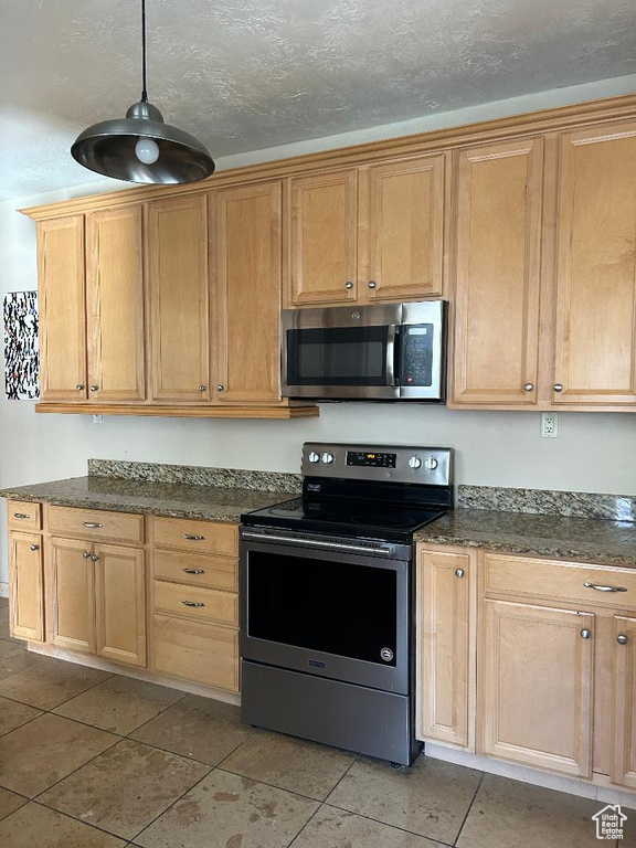 Kitchen with stainless steel appliances, light tile patterned floors, hanging light fixtures, and dark stone counters