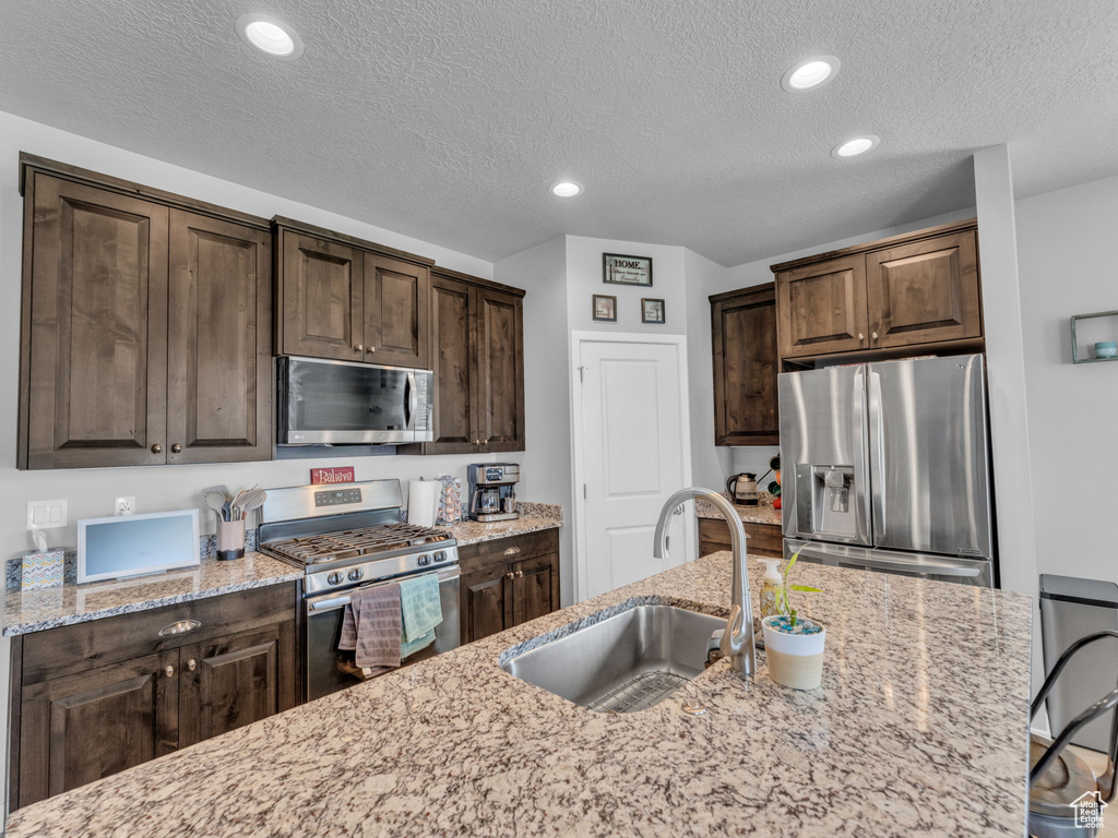 Kitchen featuring appliances with stainless steel finishes, a sink, dark brown cabinetry, a textured ceiling, and light stone counters