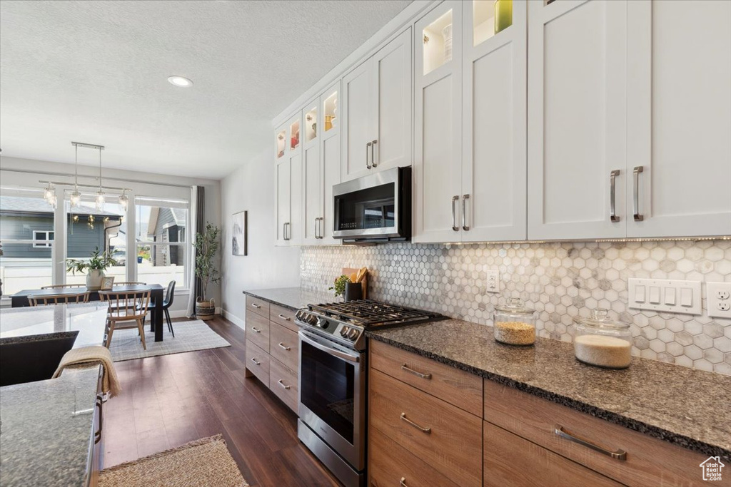Kitchen with stainless steel appliances, dark wood-type flooring, tasteful backsplash, glass insert cabinets, and dark stone countertops