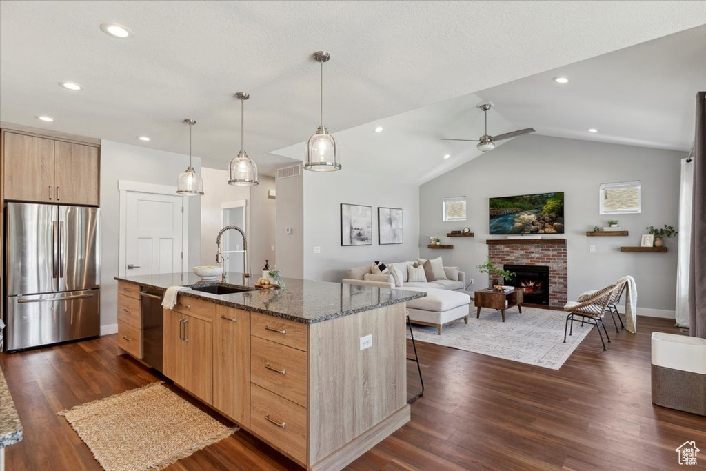Kitchen with stainless steel appliances, light brown cabinets, dark stone counters, a kitchen island with sink, and dark wood-style flooring