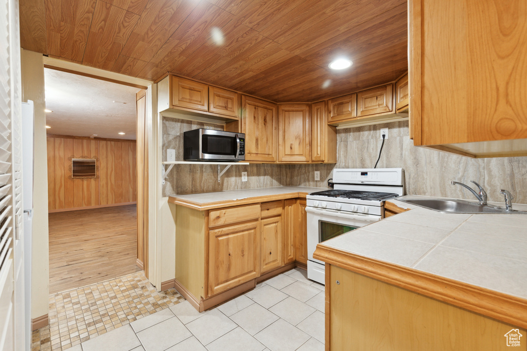 Kitchen with white range with gas stovetop, stainless steel microwave, a sink, wood ceiling, and tile counters