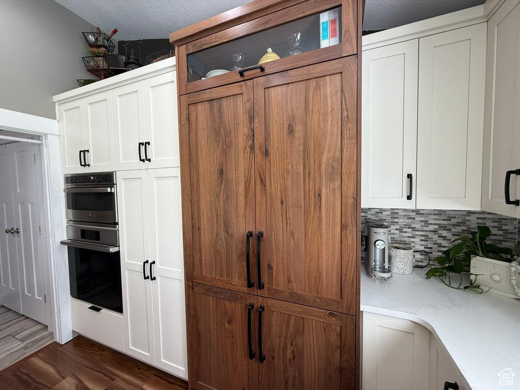 Kitchen with stainless steel double oven, tasteful backsplash, dark wood-style floors, white cabinets, and light stone counters