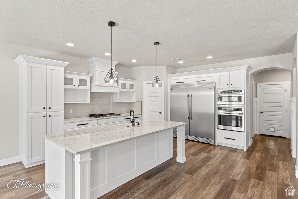 Kitchen with appliances with stainless steel finishes, a sink, dark wood-style flooring, white cabinets, and a kitchen island with sink