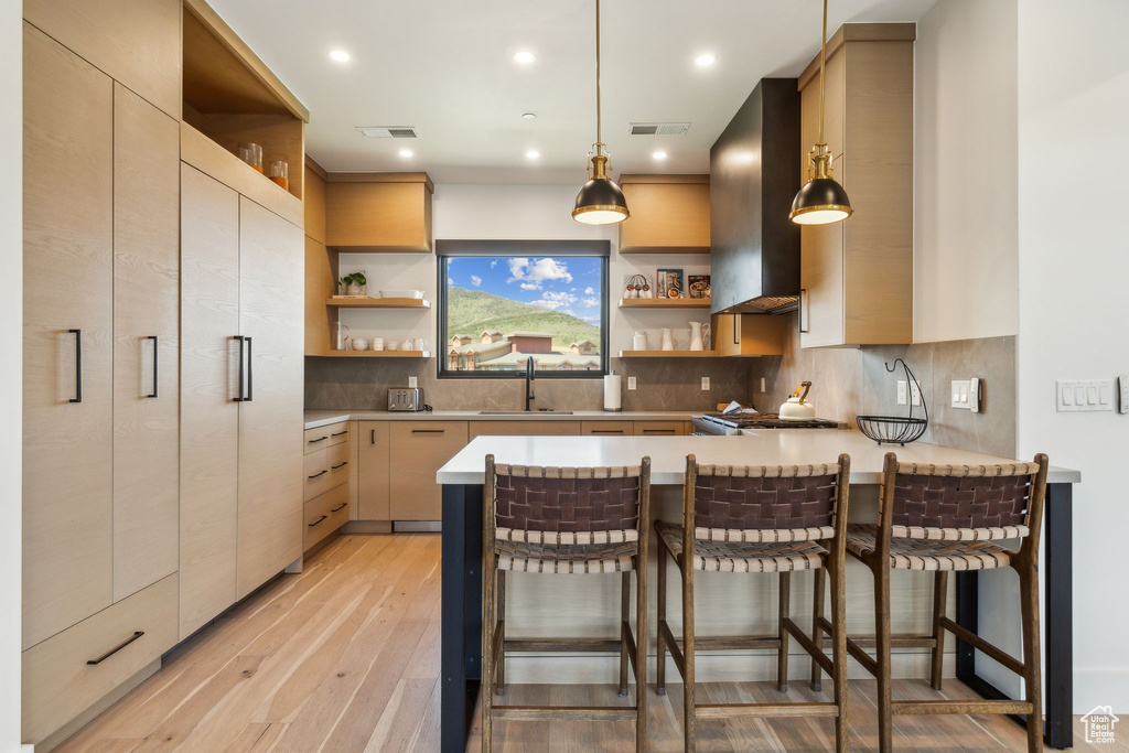 Kitchen with open shelves, a sink, a peninsula, wall chimney range hood, and light wood-type flooring